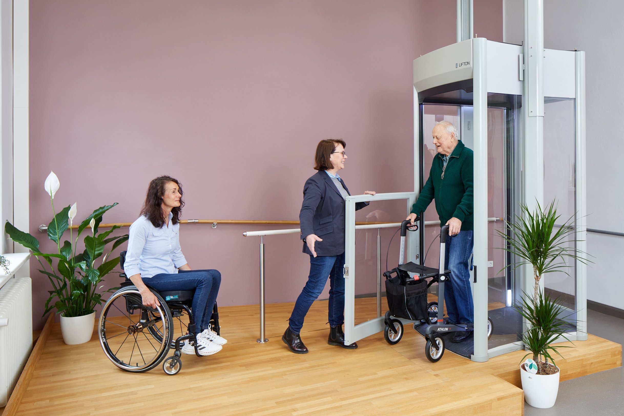 Das Bild zeigt einen Ausstellungsraum mit einem Homelift. Aus dem Lift steigt ein Besucher mit Rollator. Vor dem Lift steht eine Frau im Rollstuhl.