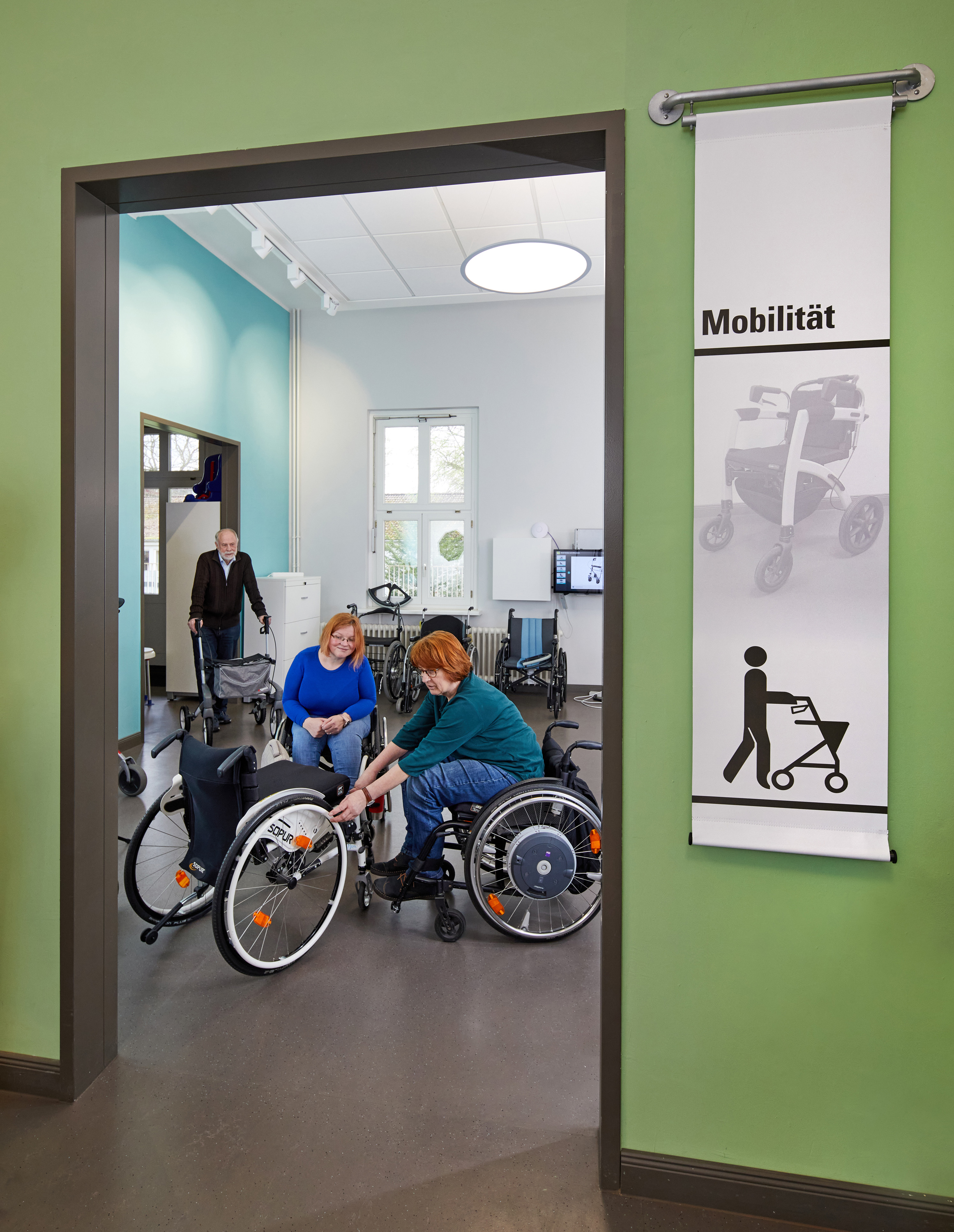 Zu sehen ist der Ausstellungsraum Mobilität. Zwei Frauen beschäftigten sich mit einem Rollstuhl und ein Mann kommt mit einem Rollator dazu.