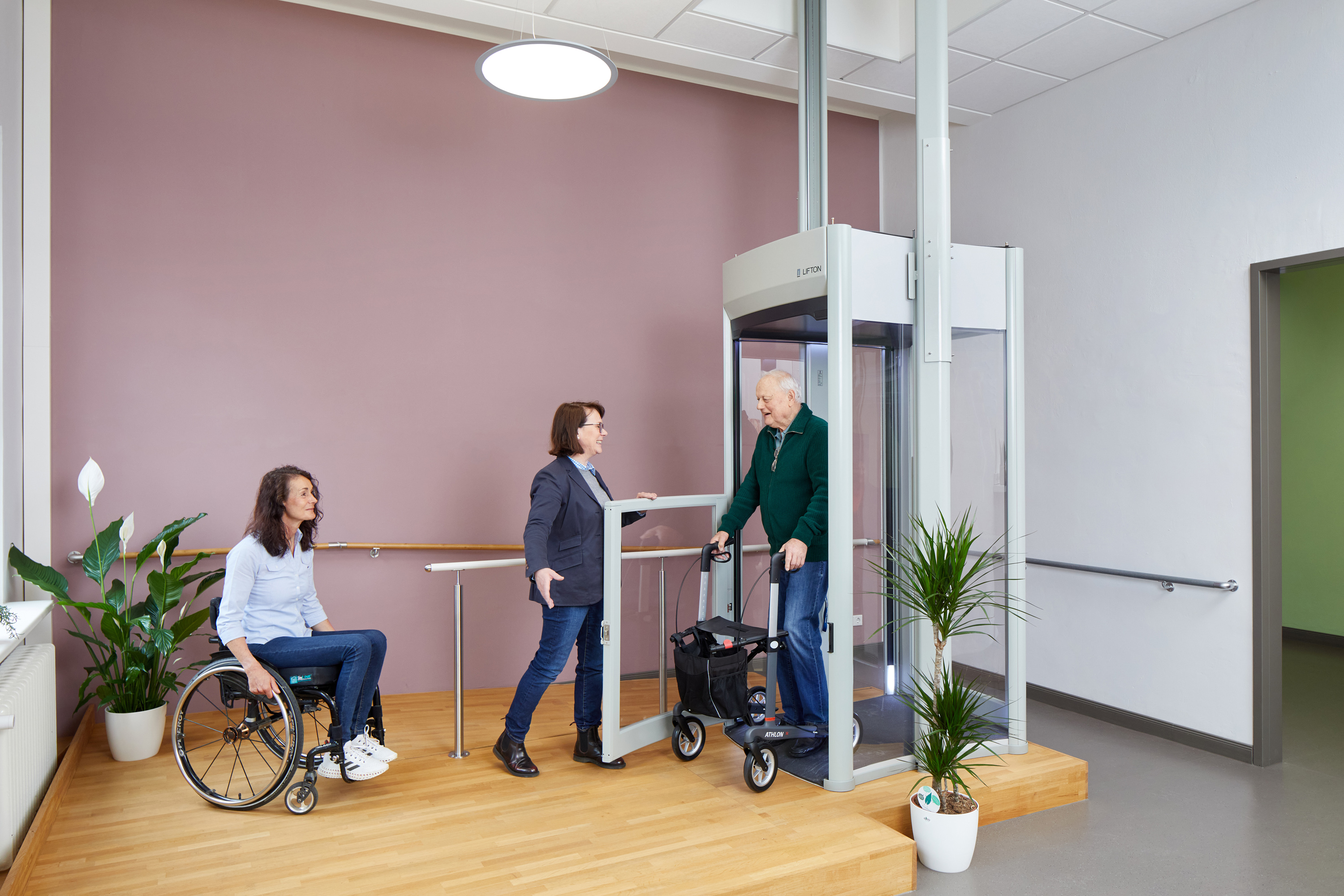 Das Bild zeigt einen Ausstellungsraum mit einem Homelift. Aus dem Lift steigt ein Besucher mit Rollator. Vor dem Lift steht eine Frau im Rollstuhl.