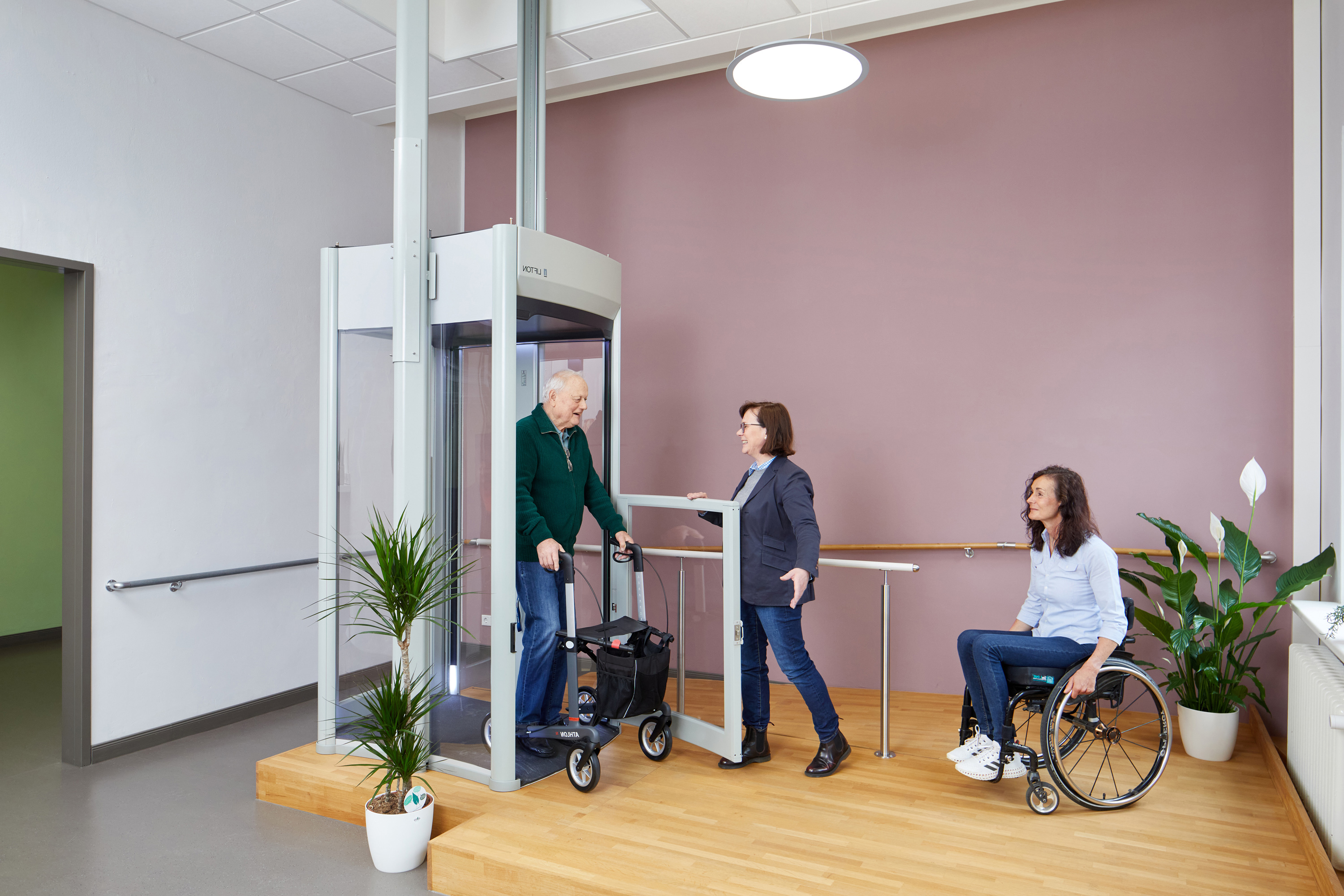 •	Das Bild zeigt einen Ausstellungsraum mit einem Homelift. Aus dem Lift steigt ein Besucher mit Rollator. Vor dem Lift steht eine Frau im Rollstuhl.