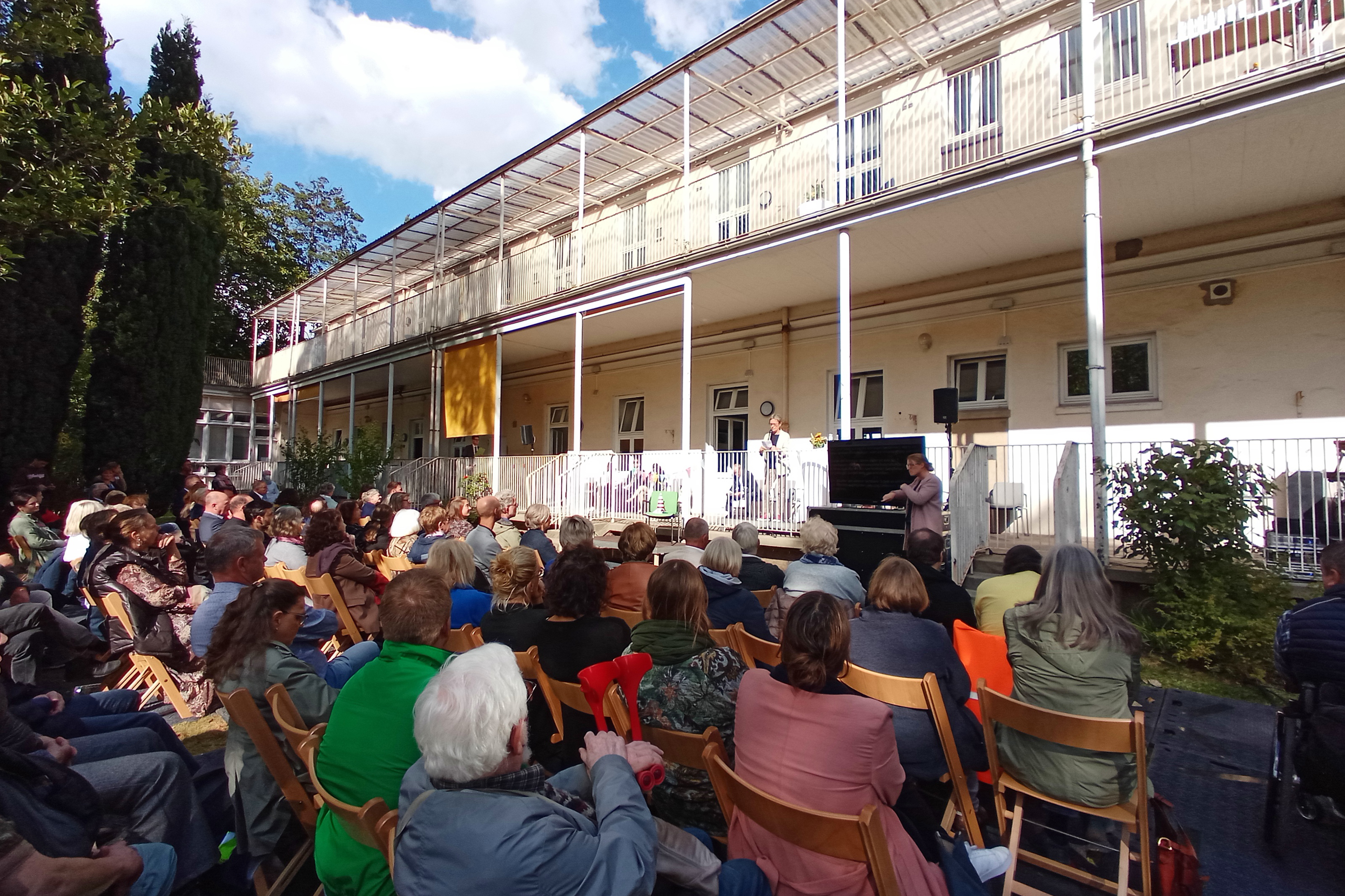 Das Bild zeigt die Rückseite des Hauses für Barrierefreiheit. Im Garten sitzen zahlreiche Teilnehmer einer Veranstaltung.