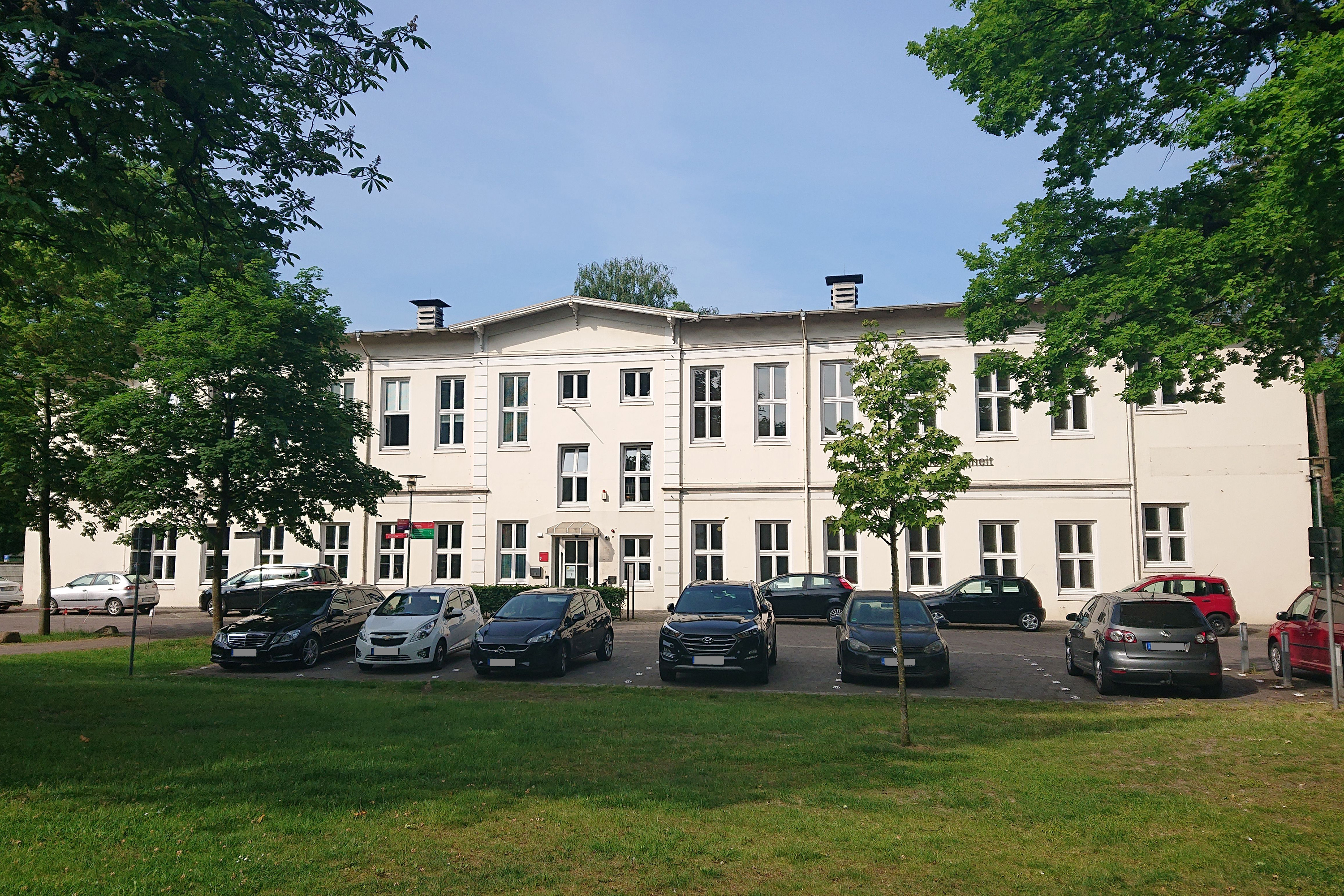 Das Bild zeigt die Vorderseite vom Haus für Barrierefreiheit und den davor befindlichen Parkplatz mit Bäumen.